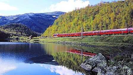 Een rode trein rijdt langs een fjord. Het is helder weer, de groene rotswand en het rood van de trein worden weerspiegeld in het water. 